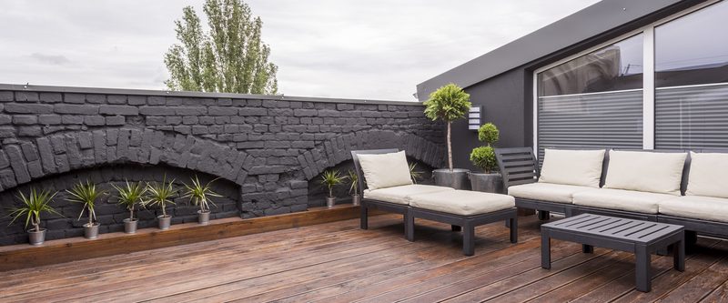 Panorama view of modern rooftop terrace with dark wood deck flooring, plants, brick fence and black garden furniture P