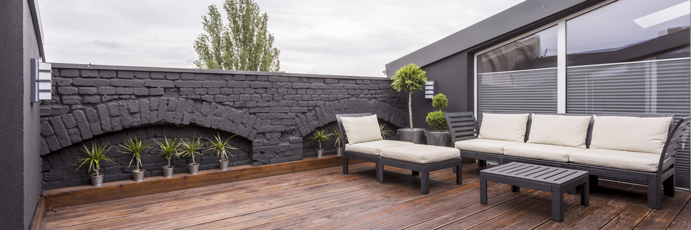 Panorama view of modern rooftop terrace with dark wood deck flooring, plants, brick fence and black garden furniture P
